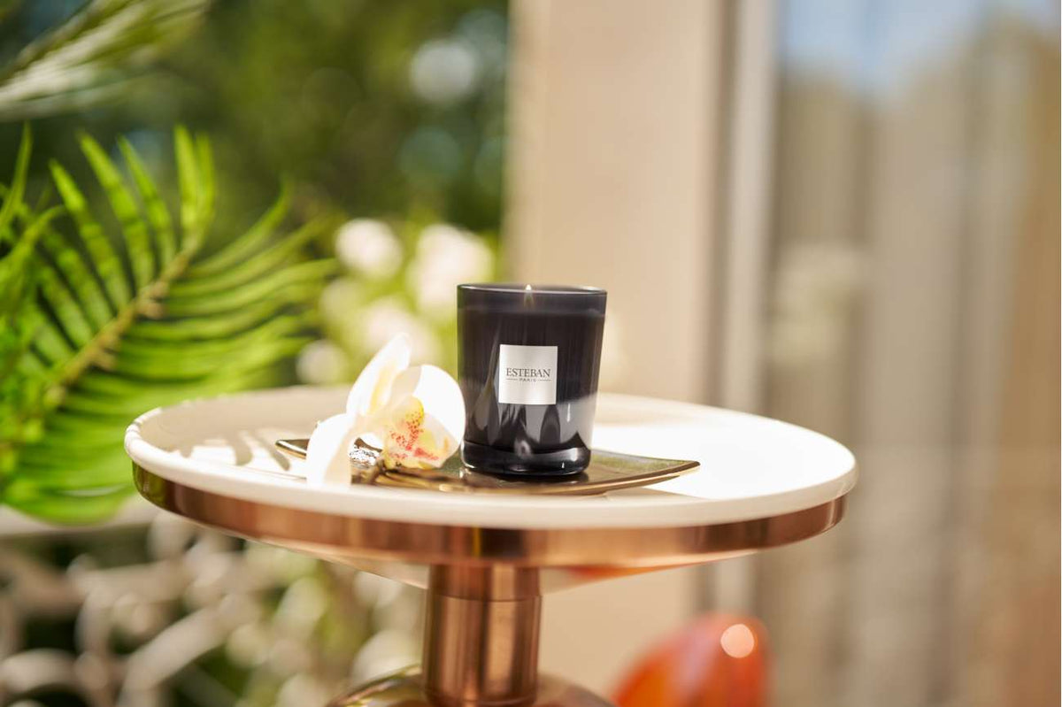 Bougie parfumée Esteban Paris Ambre & Vanille Étoilée en verre fumé noir, présentée sur un plateau doré avec une orchidée blanche dans une ambiance lumineuse et végétale