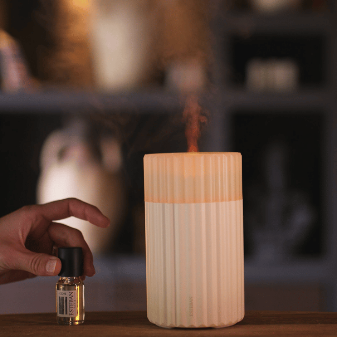 Diffuseur électrique de parfum Esteban Paris en céramique blanche nervurée, diffusant une fine brume parfumée, présenté avec une huile parfumée Cèdre dans une ambiance intérieure chaleureuse
